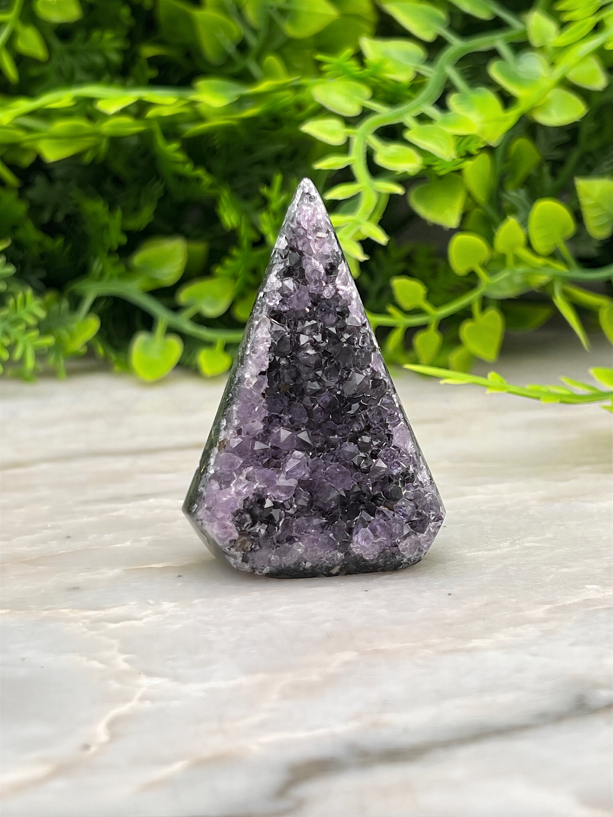 A triangular piece of purple amethyst crystal sits on a marble surface, surrounded by lush green foliage.