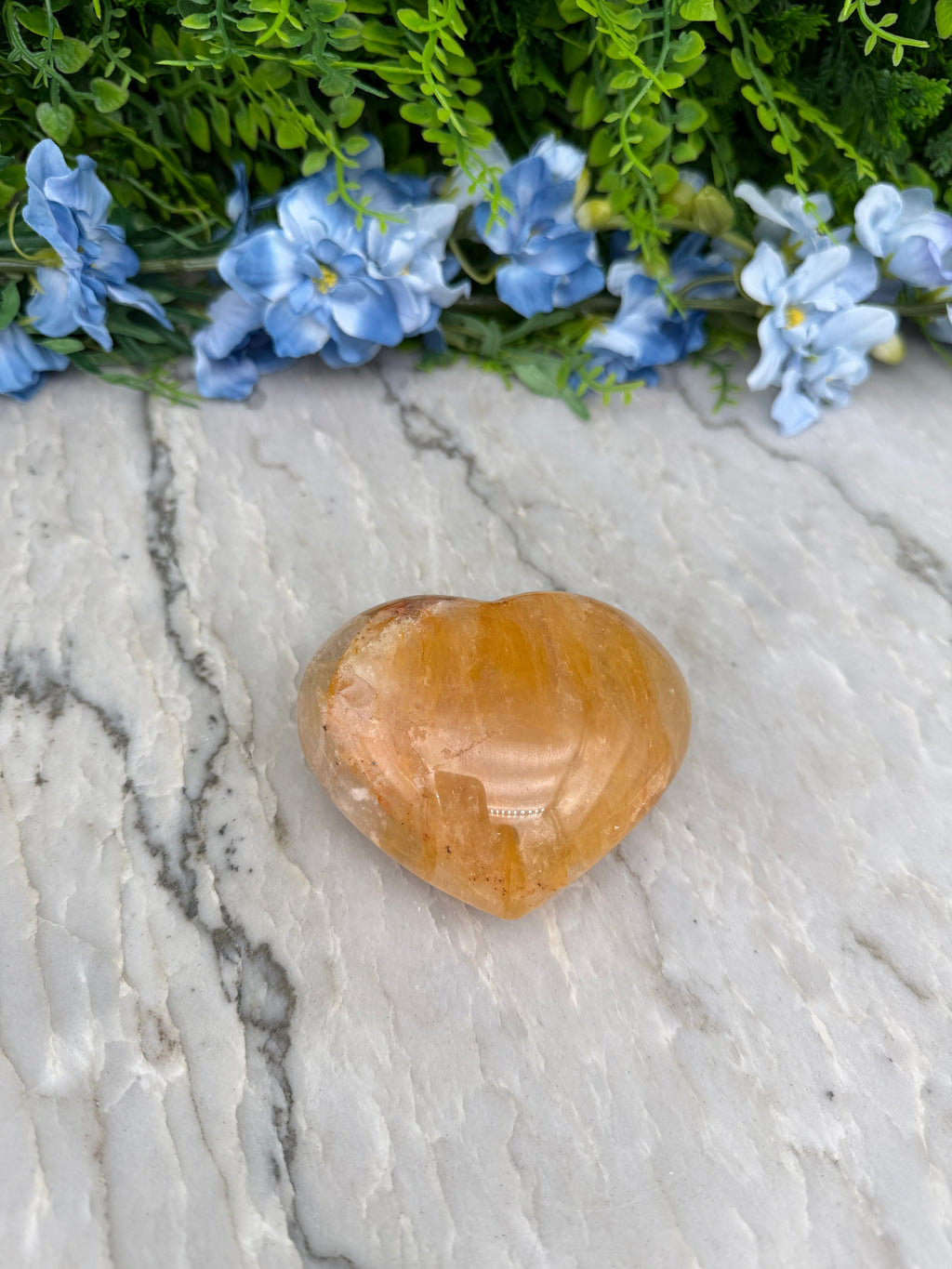 a heart-shaped amber gemstone resting on a marble surface, surrounded by blue flowers.