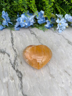 a heart-shaped amber gemstone resting on a marble surface, surrounded by blue flowers.