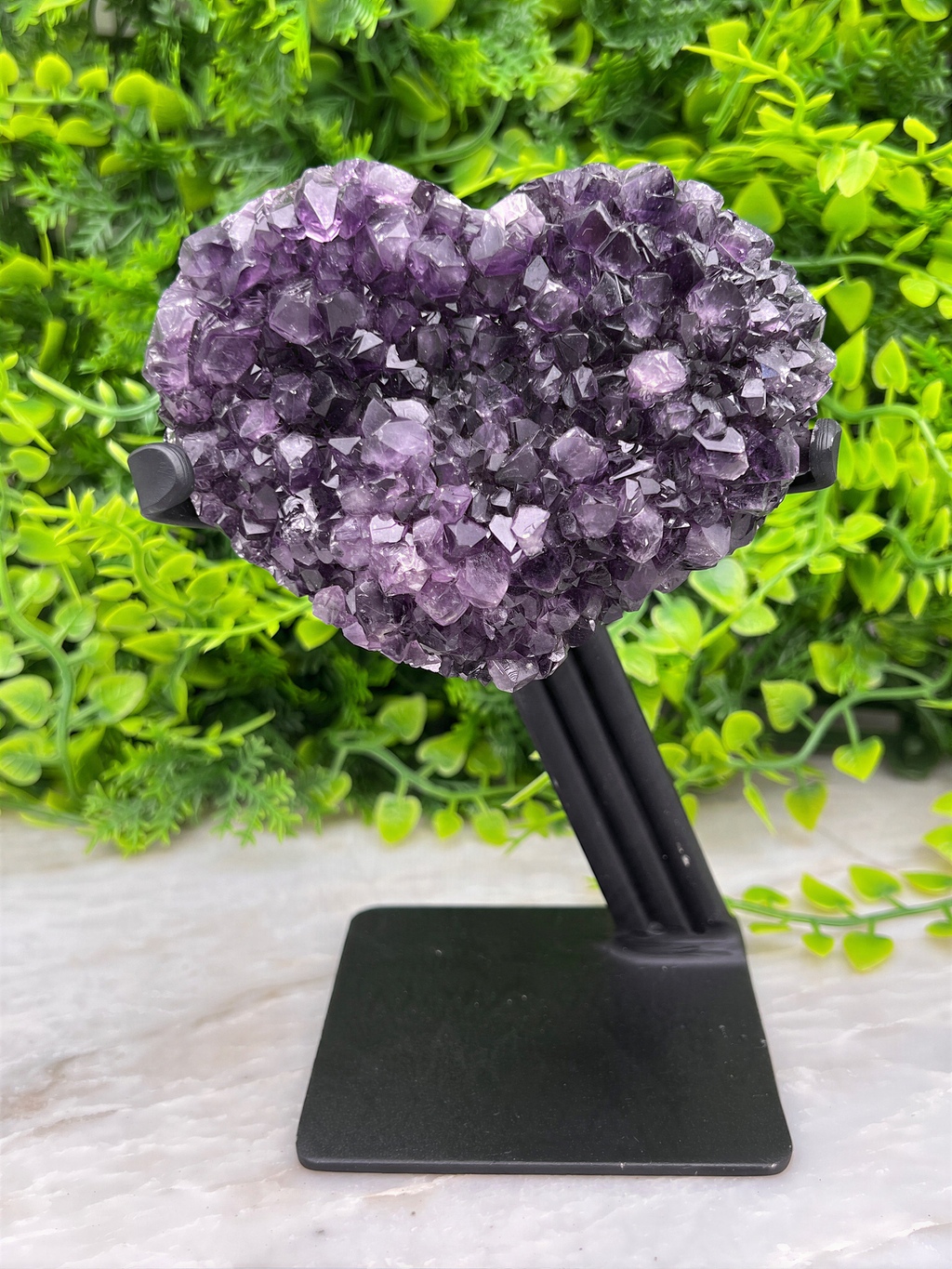 A heart-shaped crystal formation made of purple amethyst crystals is displayed on a black stand, surrounded by green foliage.