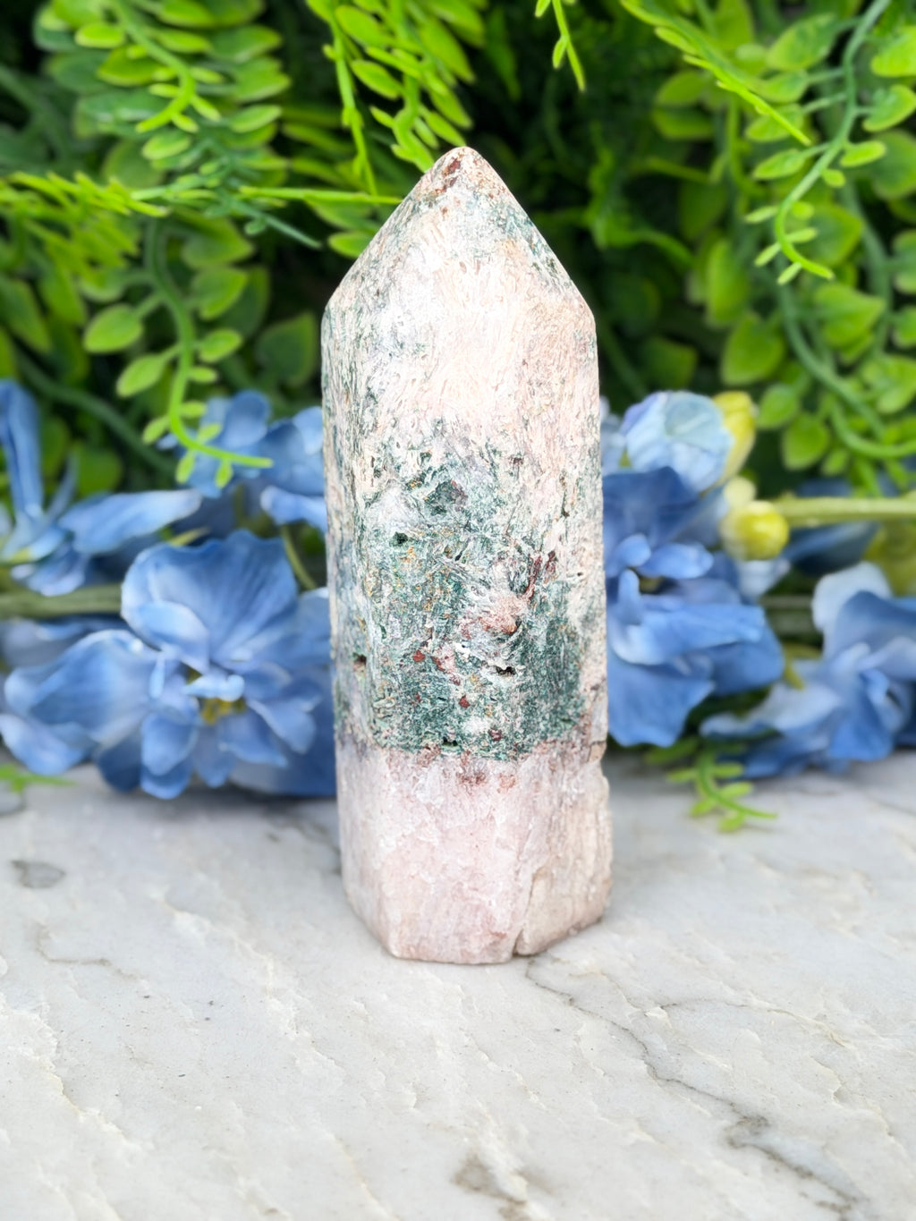 A light pink crystal with green and brown speckles sits on a white marble surface, surrounded by blue flowers.