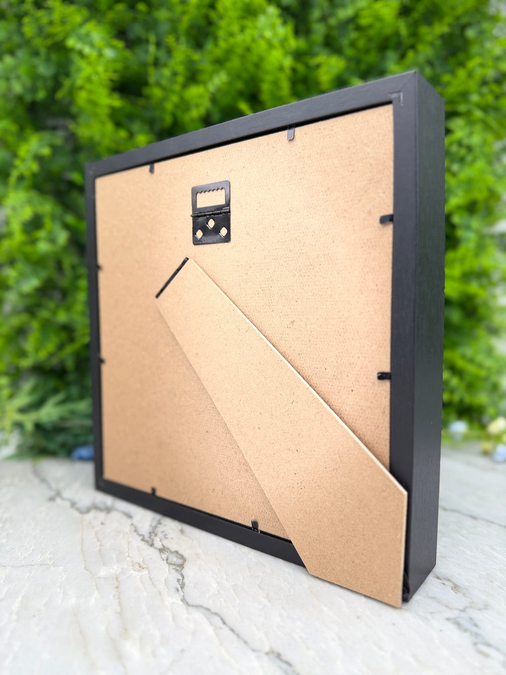 a square picture frame with a black border, placed on a white surface against a background of green foliage.
