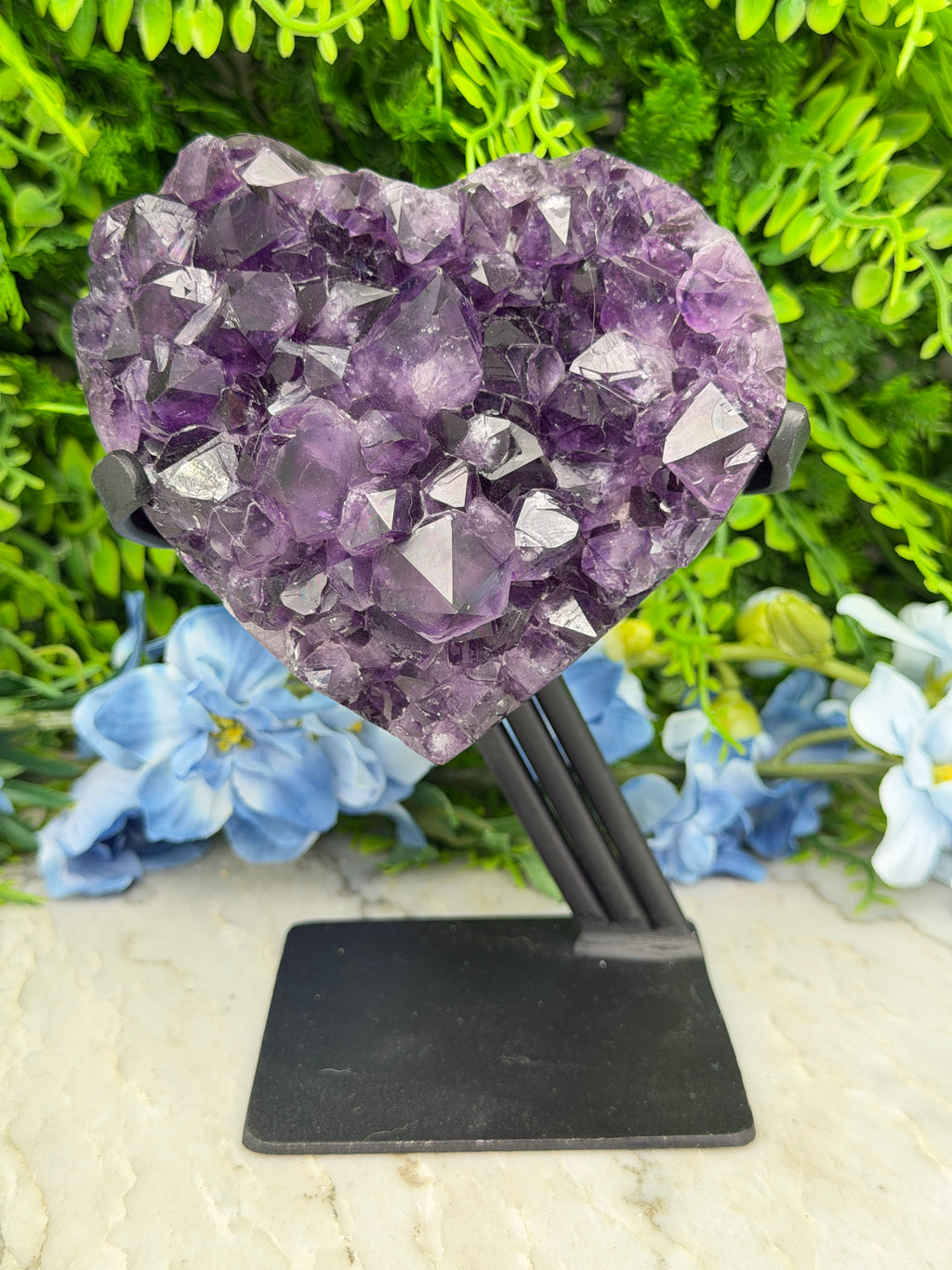 A heart-shaped amethyst crystal is mounted on a black metal bracket, set against a backdrop of lush green foliage and blue flowers.