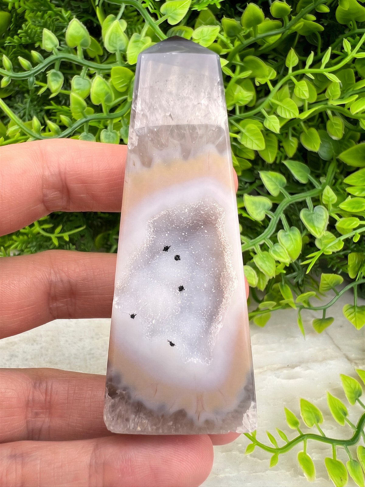 A hand is holding a clear geode with a unique rock formation, set against a backdrop of green foliage.