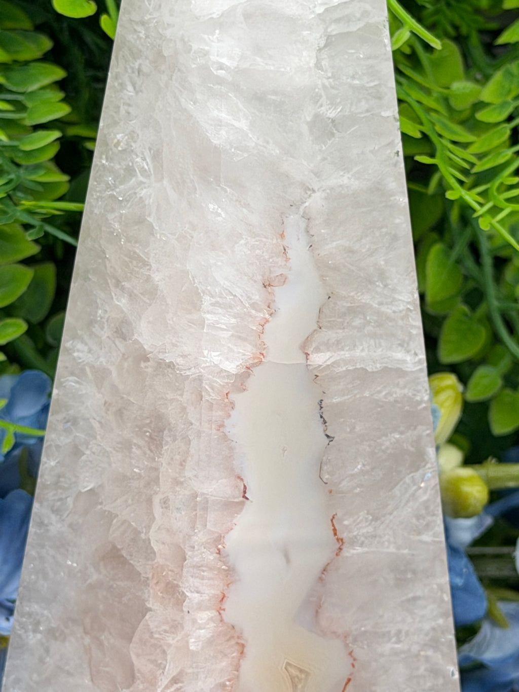 a close-up view of a white crystal or stone with a unique, jagged pattern. The crystal is set against a background of green leaves and flowers, creating a contrast between the natural and man-made elements.