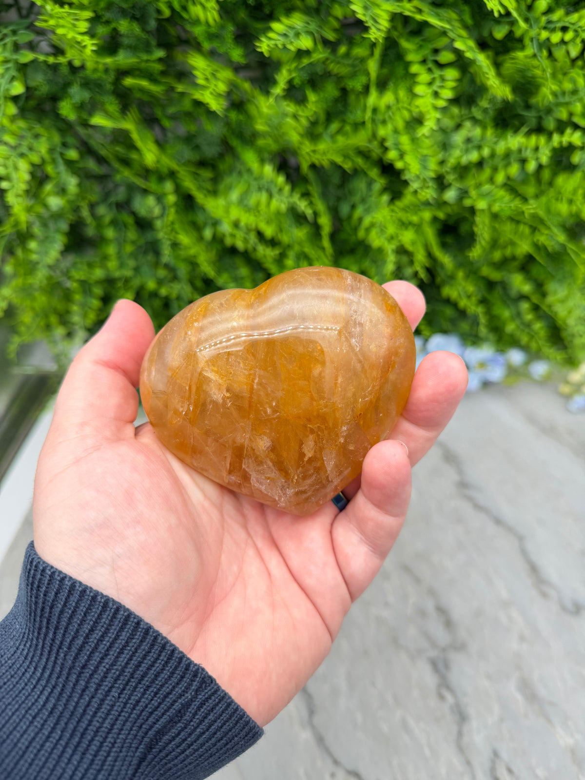 A hand holding a yellowish-brown crystal or gemstone against a backdrop of green foliage.
