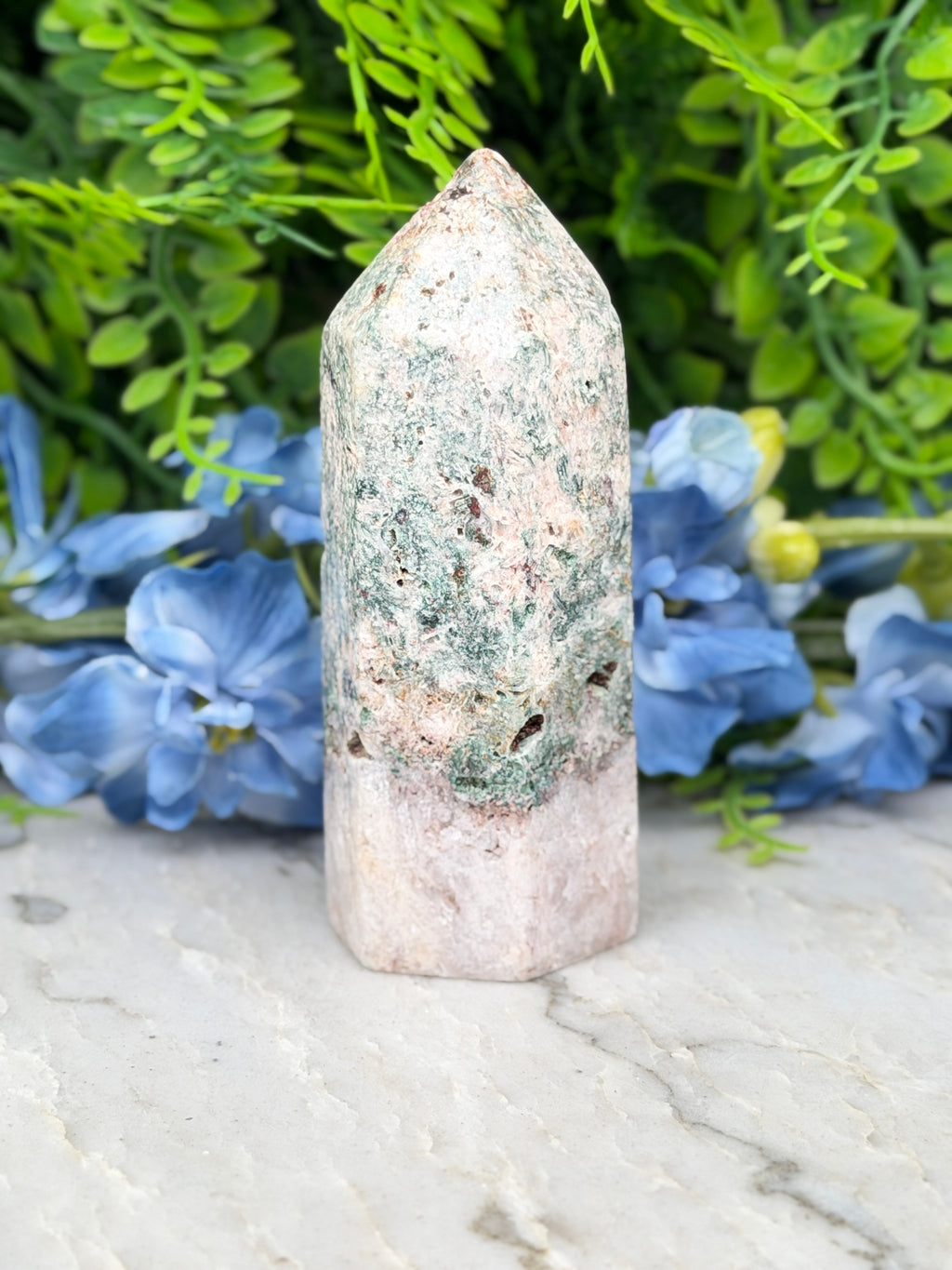 A white and green crystal sits on a marble surface, surrounded by blue flowers.