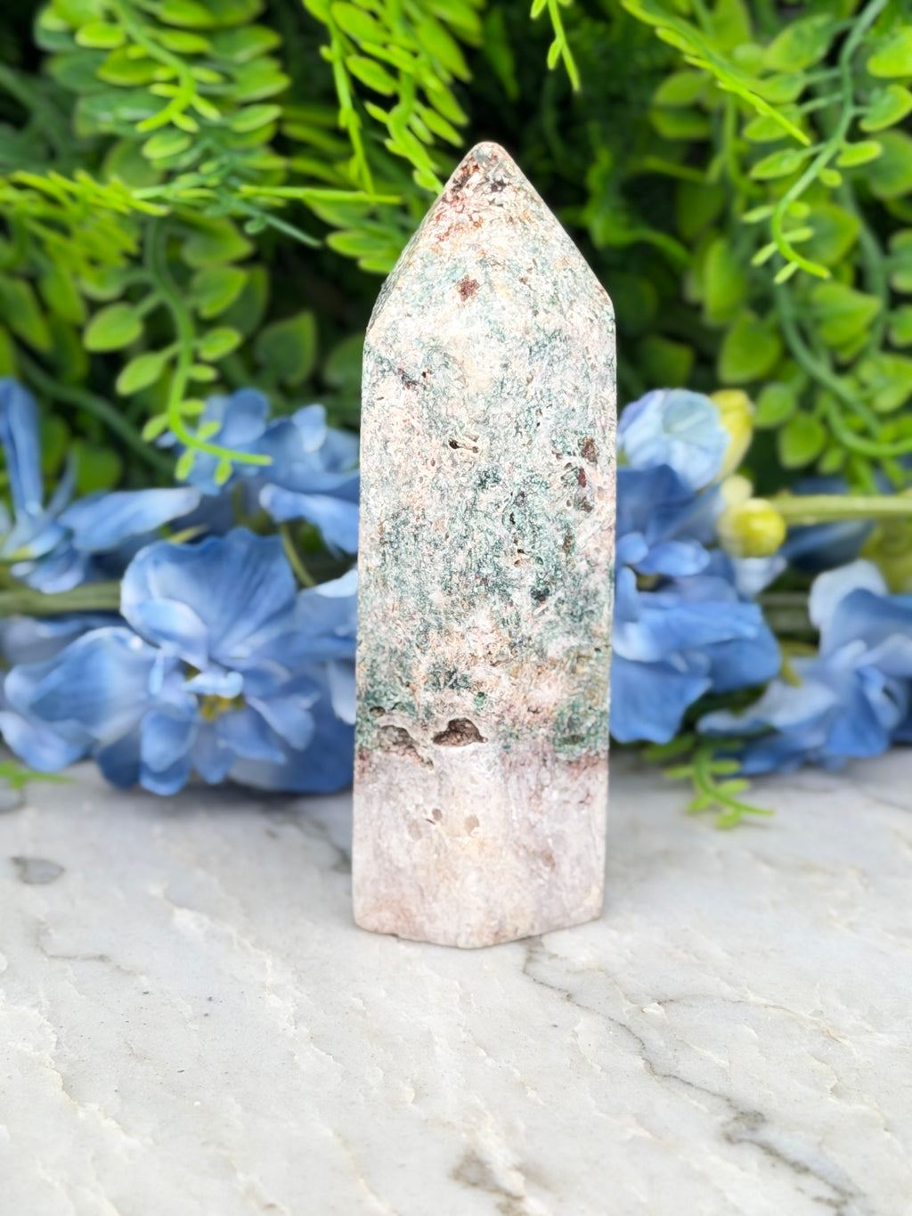 A white and green crystal sits on a marble surface, surrounded by blue flowers.