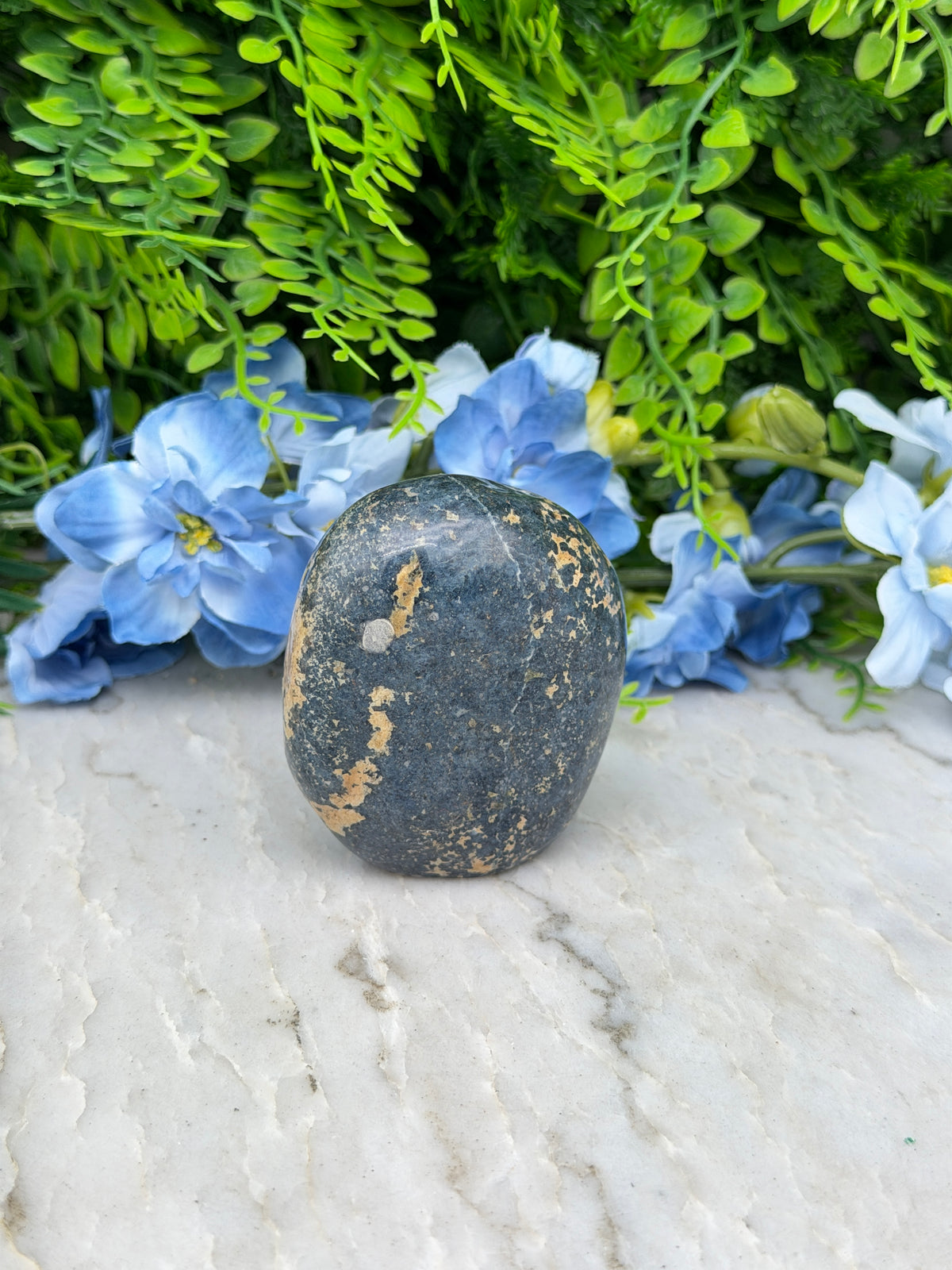 A black rock with a speckled surface rests on a white surface, surrounded by vibrant blue flowers.