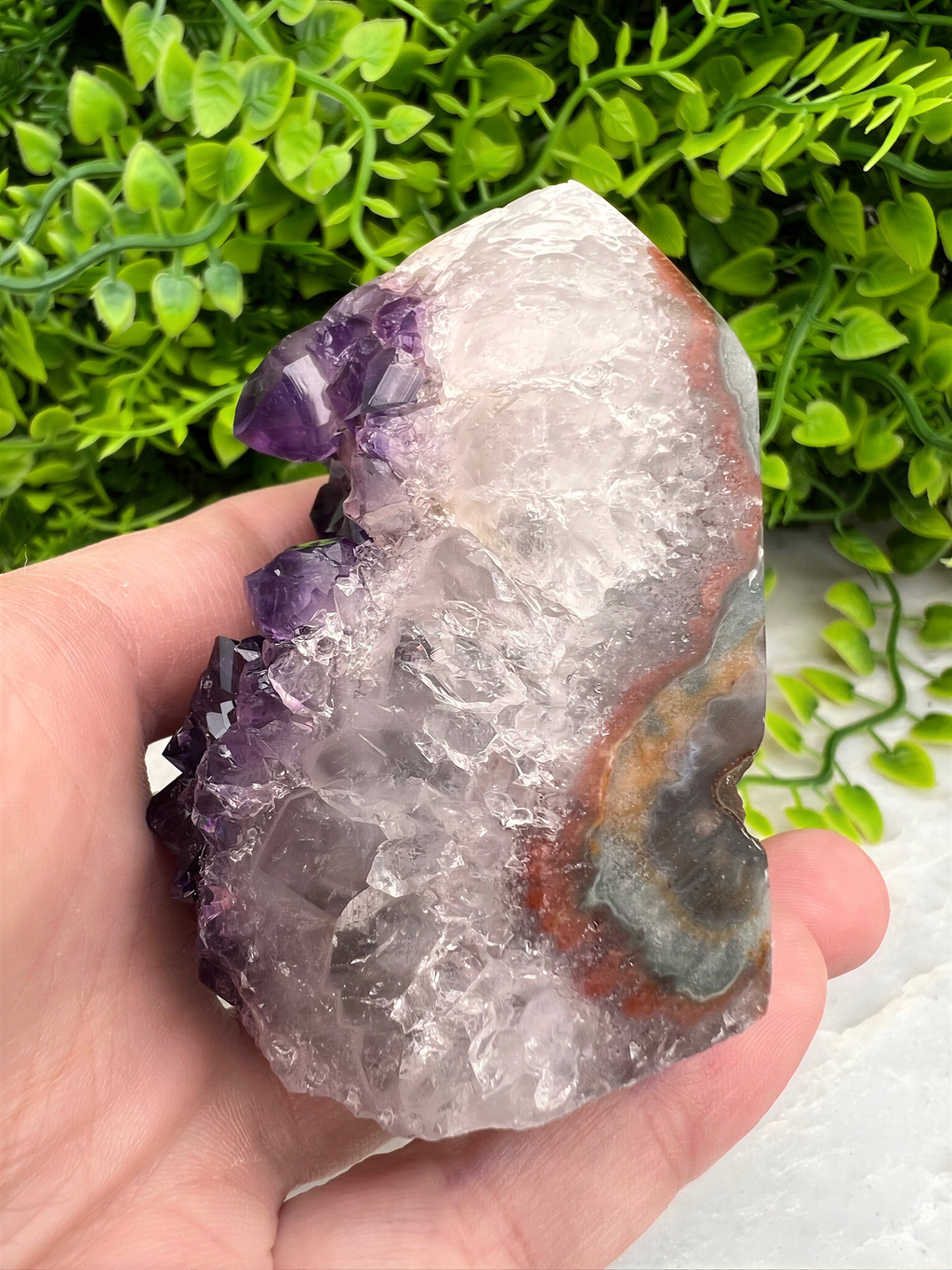 A hand holding a piece of amethyst crystal against a backdrop of green foliage.
