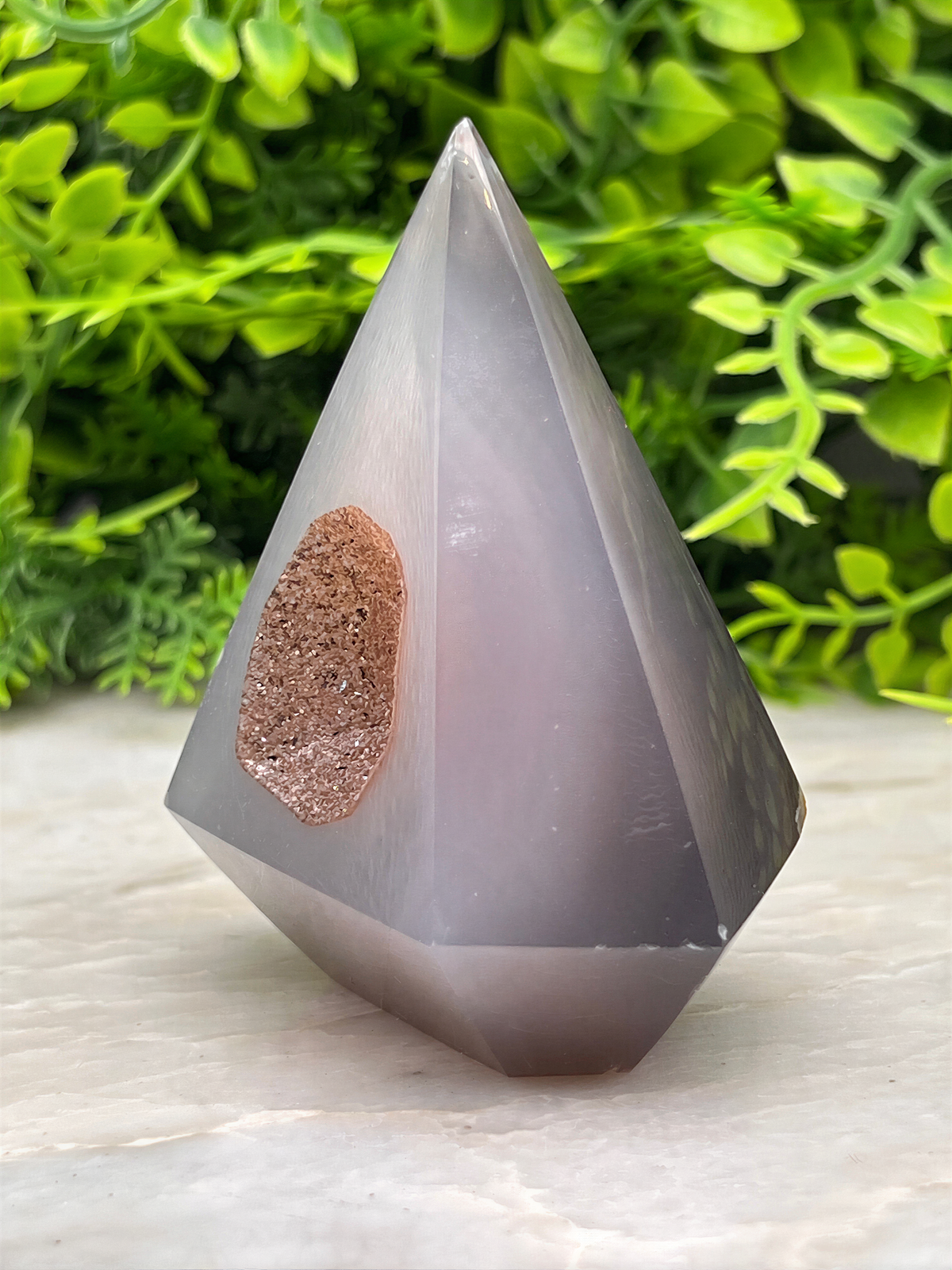 A gray, triangular crystal sits on a white surface, with a small piece of brown rock attached to it.