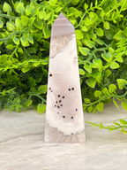 A pink and white crystal formation with black spots is displayed on a white surface, surrounded by green foliage.