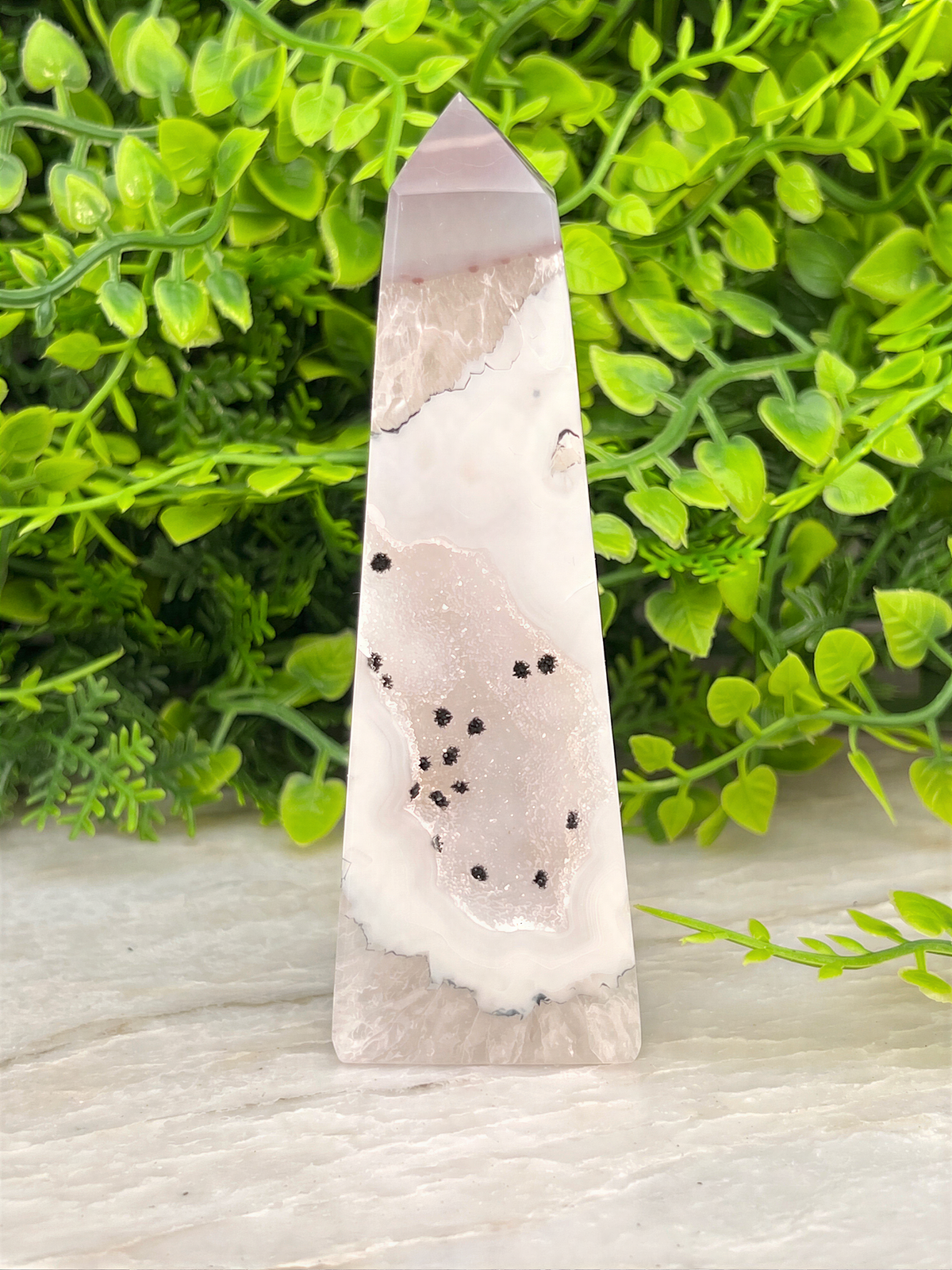 A pink and white crystal formation with black spots is displayed on a white surface, surrounded by green foliage.