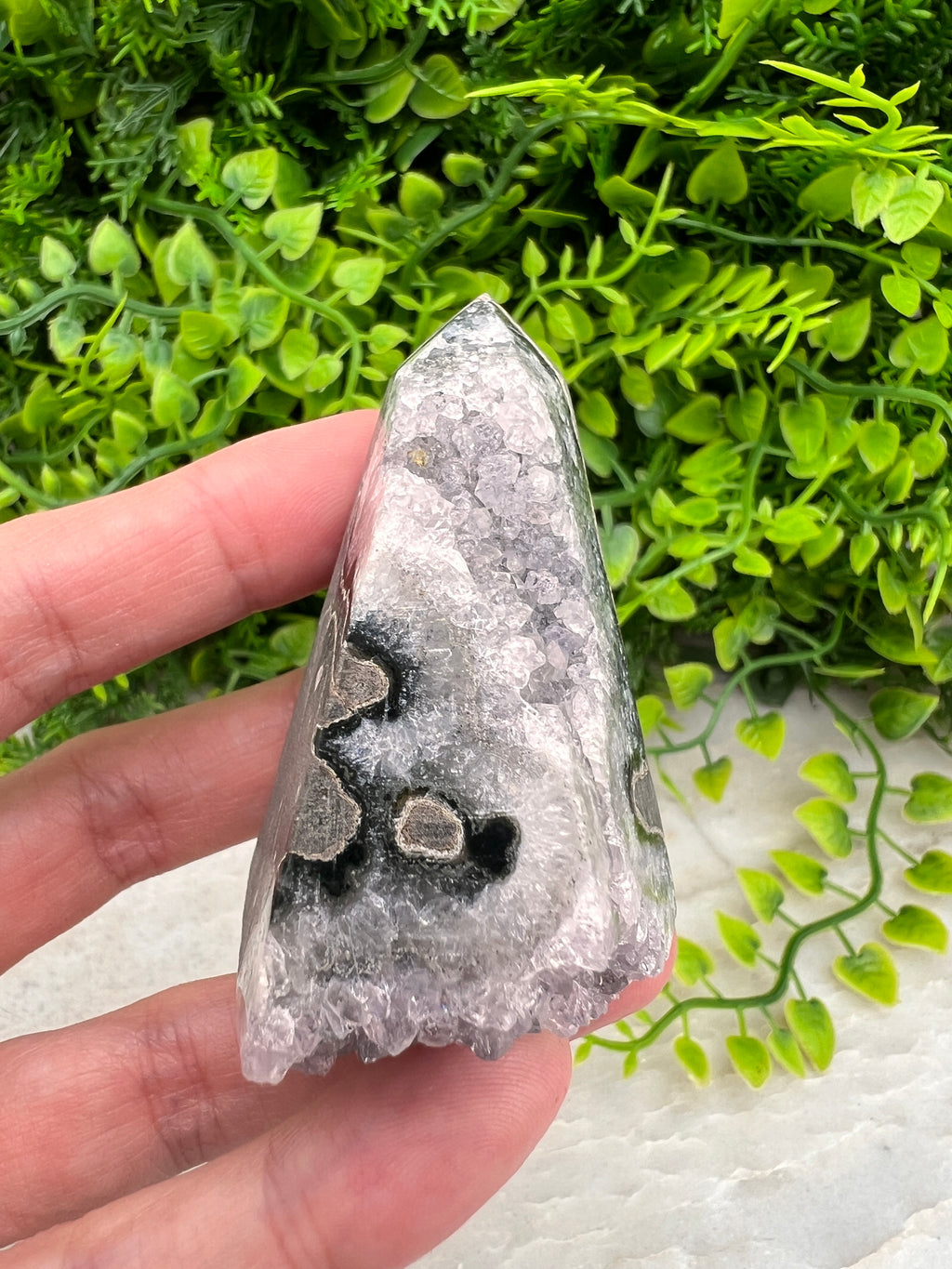A hand is holding a piece of a crystal or gemstone, with a background of green plants.