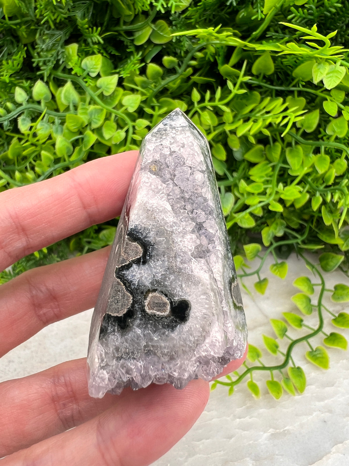 A hand is holding a piece of a crystal or gemstone, with a background of green plants.