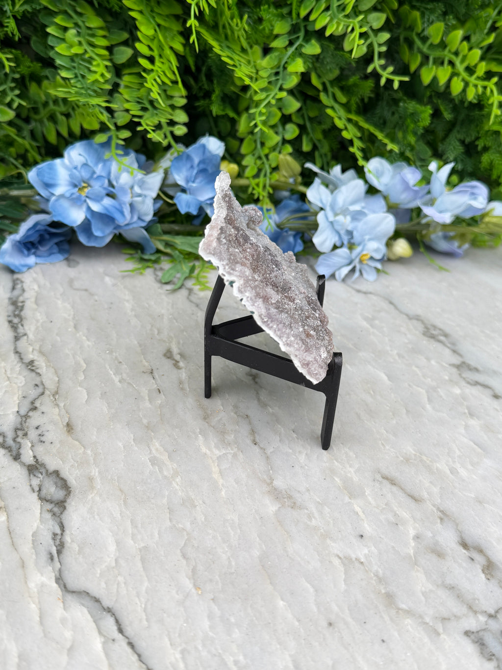 a piece of amethyst crystal placed on a black stand, surrounded by blue flowers and greenery.