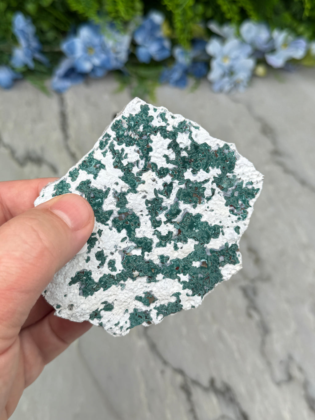 A hand is holding a piece of rock with a speckled surface, against a background of blue flowers.