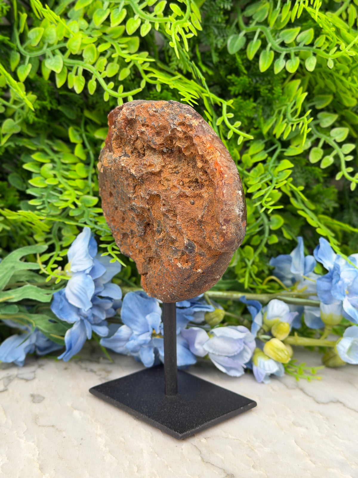 a large, irregularly shaped rock or stone on display, placed on a black stand in front of a backdrop of green foliage and blue flowers.