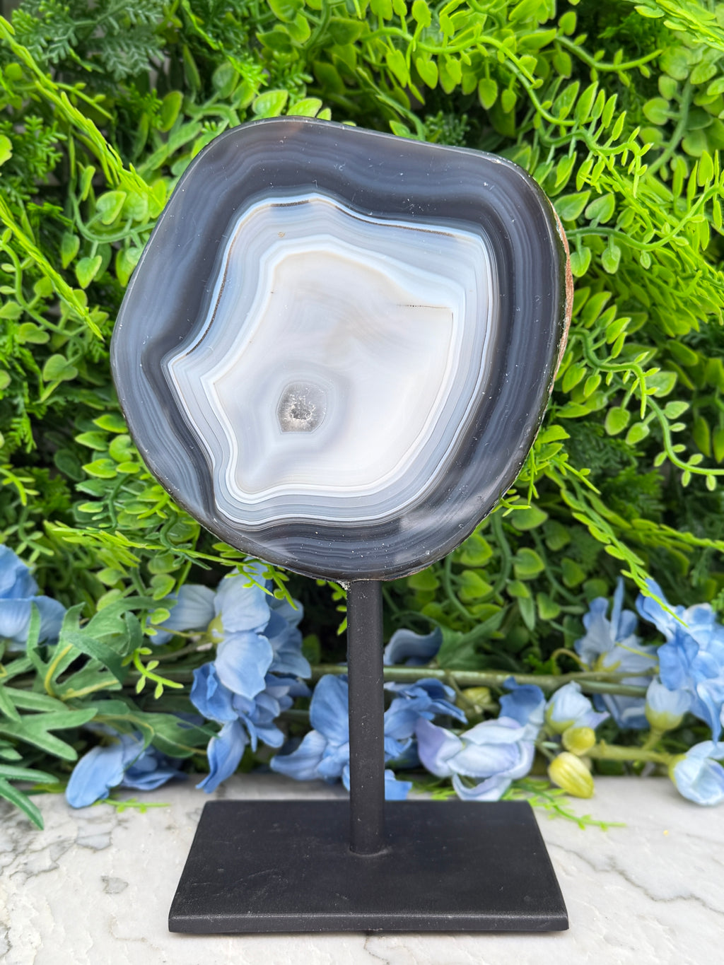 a black rock slice with a white and gray striped pattern, placed on a black stand in front of a backdrop of green foliage and blue flowers.