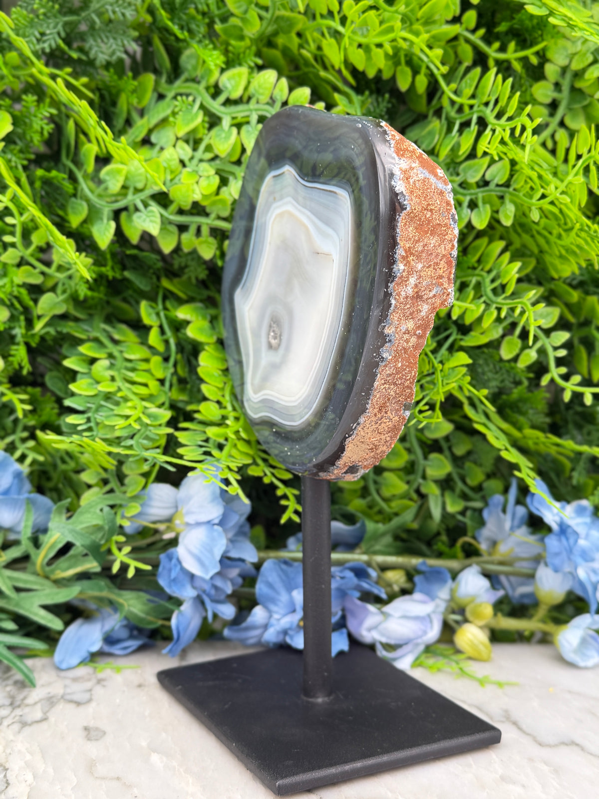a large, irregularly shaped rock with a white and black striped pattern, placed on a black stand in front of a backdrop of lush green foliage and blue flowers.