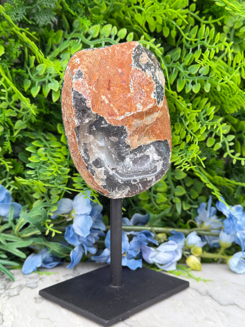 a rock with a black and orange color pattern, placed on a black stand and surrounded by blue flowers.