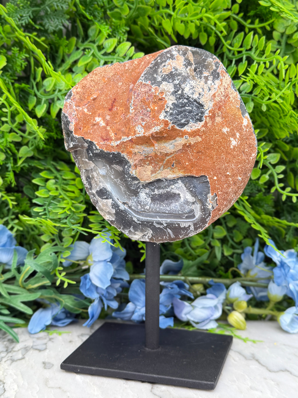 a rock with a mix of orange and black colors, placed on a black stand in front of a backdrop of green foliage and blue flowers.