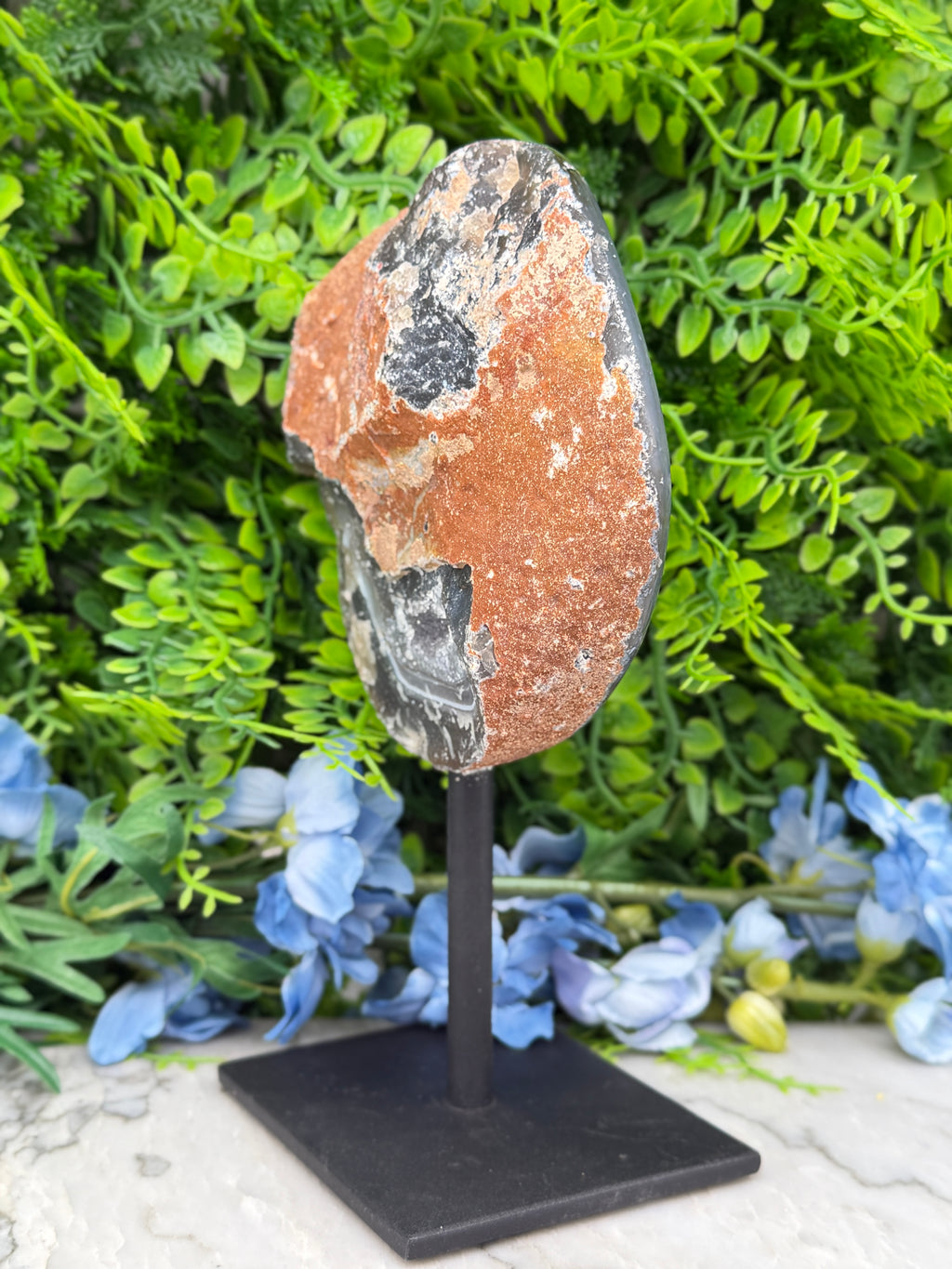 a rock formation with a black and orange color pattern, placed on a black stand in front of a backdrop of green foliage and blue flowers.