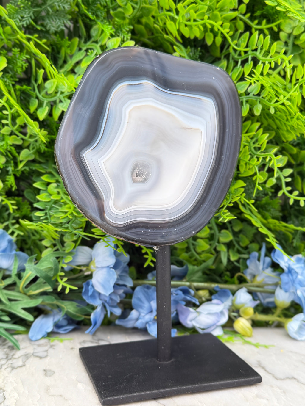 a large, black agate stone with a unique, striped pattern, displayed on a black stand in front of a backdrop of lush green foliage and blue flowers.