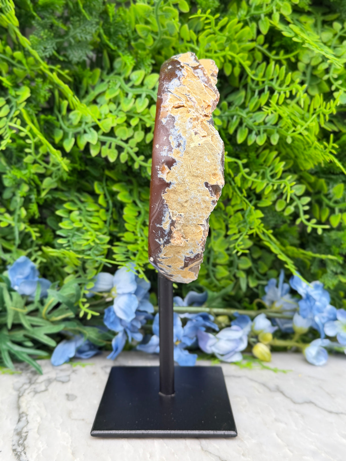 a rock formation with a mix of green and brown hues, placed on a black stand in front of a backdrop of lush green foliage and blue flowers.