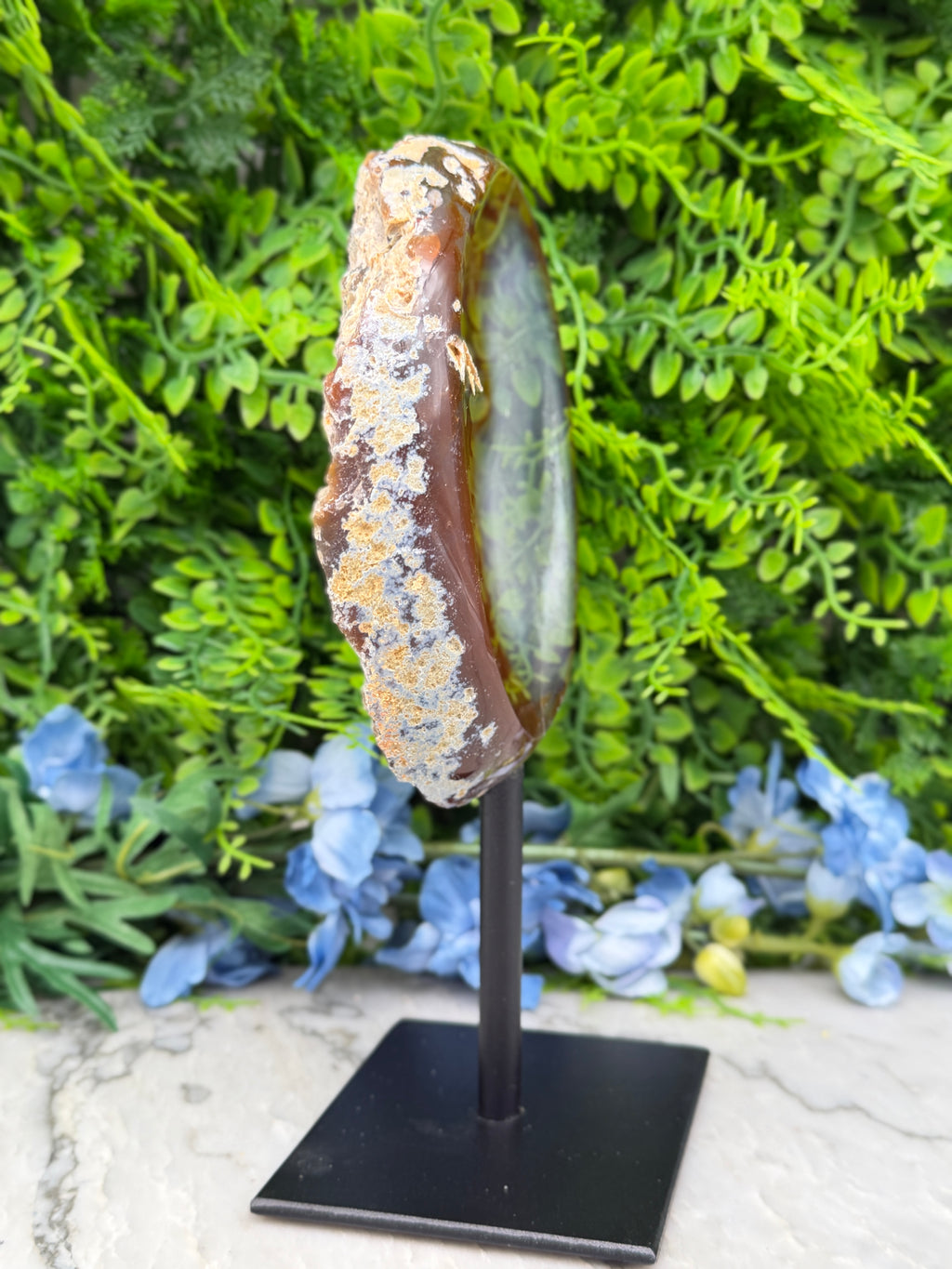 a unique, handcrafted piece of art, a wooden sculpture of a round, ornate object, possibly a piece of jewelry or a decorative item, displayed on a black stand. The sculpture is set against a backdrop of lush green foliage, creating a visually appealing contrast between the natural and man-made elements.