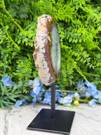 a unique, handcrafted piece of art, a wooden sculpture of a round, ornate object, possibly a piece of jewelry or a decorative item, displayed on a black stand. The sculpture is set against a backdrop of lush green foliage, creating a visually appealing contrast between the natural and man-made elements.
