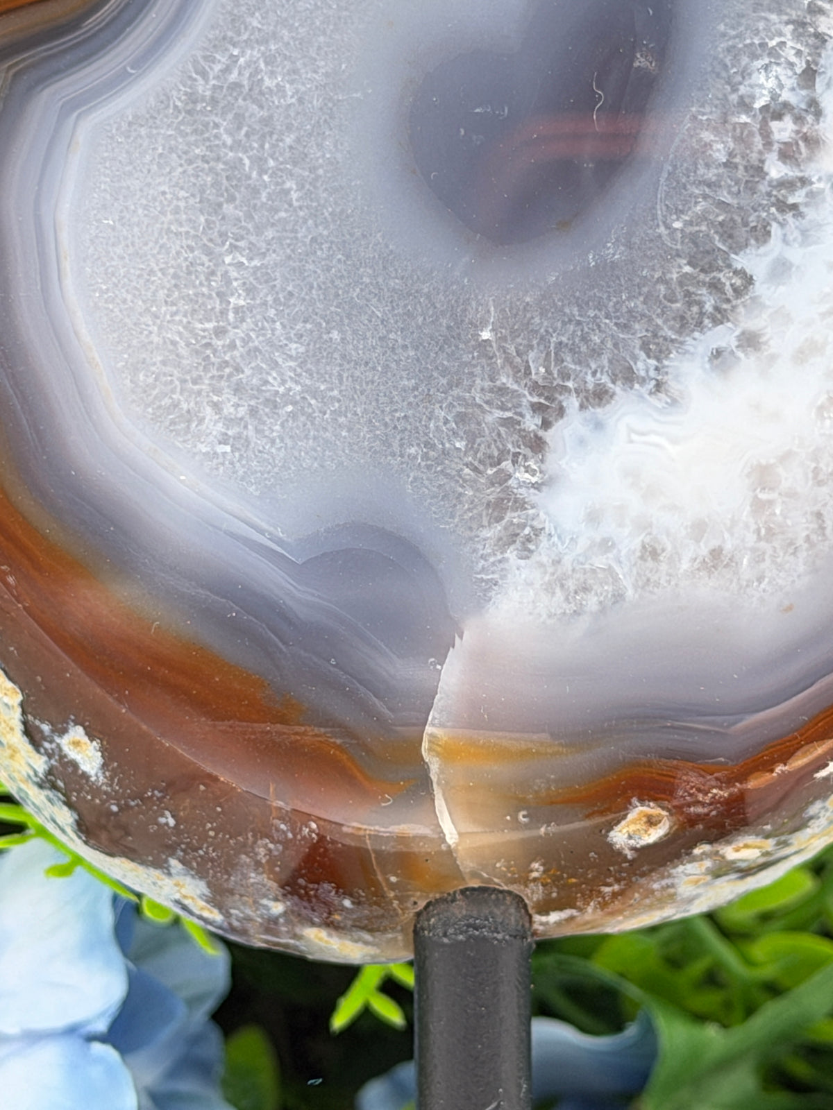 a close-up view of a natural rock formation with a unique, striped pattern. The rock appears to be a combination of white, brown, and orange colors, with a rough, textured surface. The rock is set against a backdrop of greenery, creating a contrast between the natural and man-made elements.