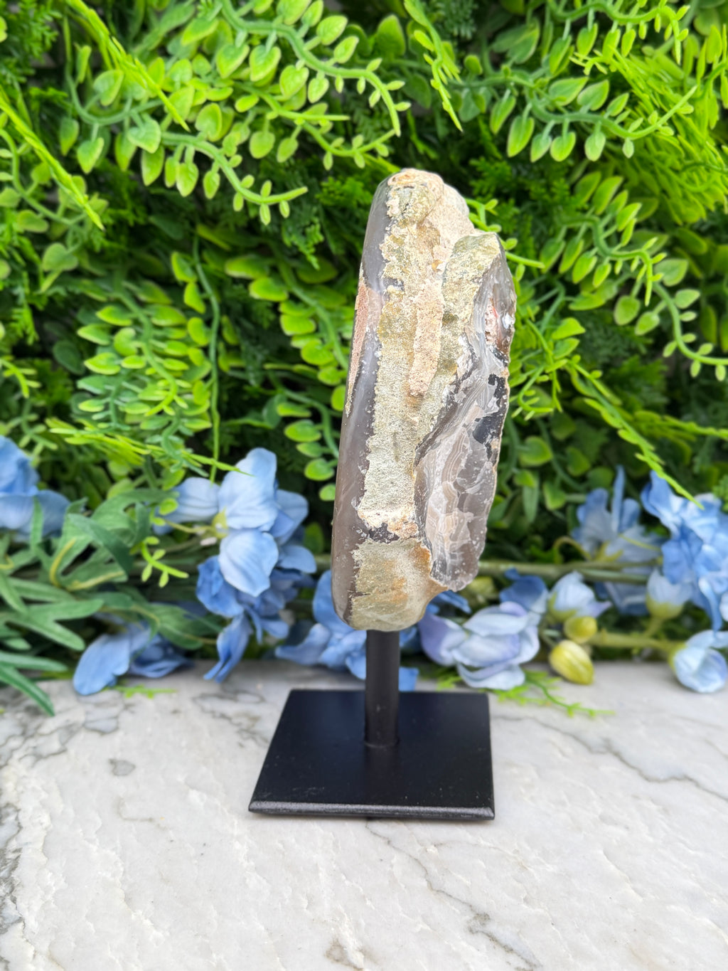 a rock formation with a small piece of rock on display, placed on a black stand in front of a backdrop of green foliage and blue flowers.