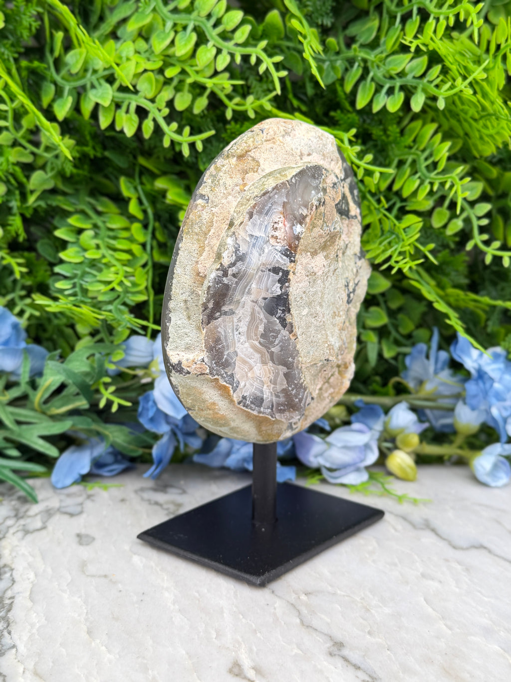 a rock formation with a unique, irregular shape, surrounded by lush green foliage and blue flowers.
