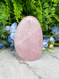 A pink amethyst crystal sits on a marble surface, surrounded by lush green foliage and delicate blue flowers.