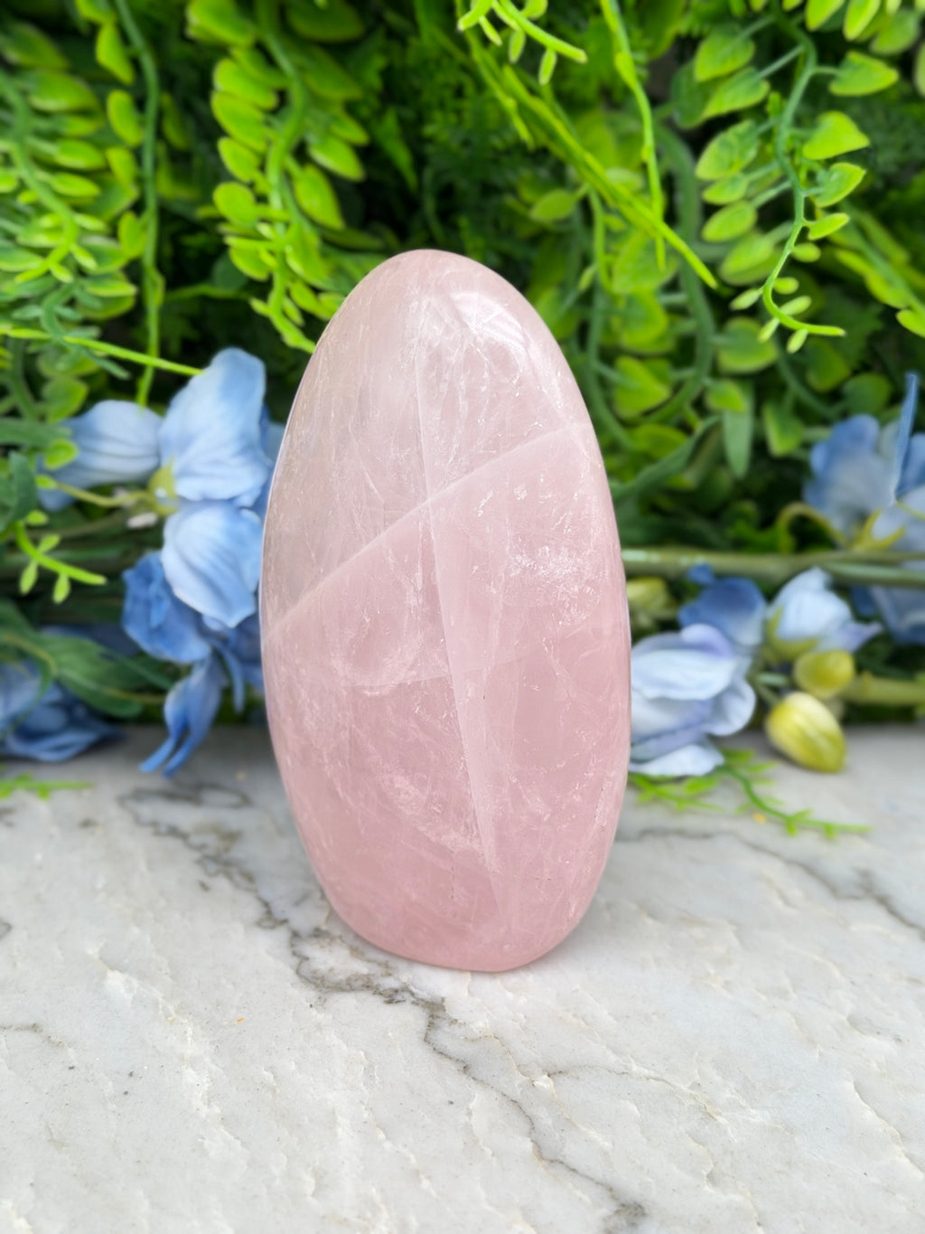 A pink amethyst stone is placed on a marble surface, surrounded by blue flowers and green foliage.