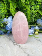 A pink amethyst stone is placed on a marble surface, surrounded by blue flowers and green foliage.