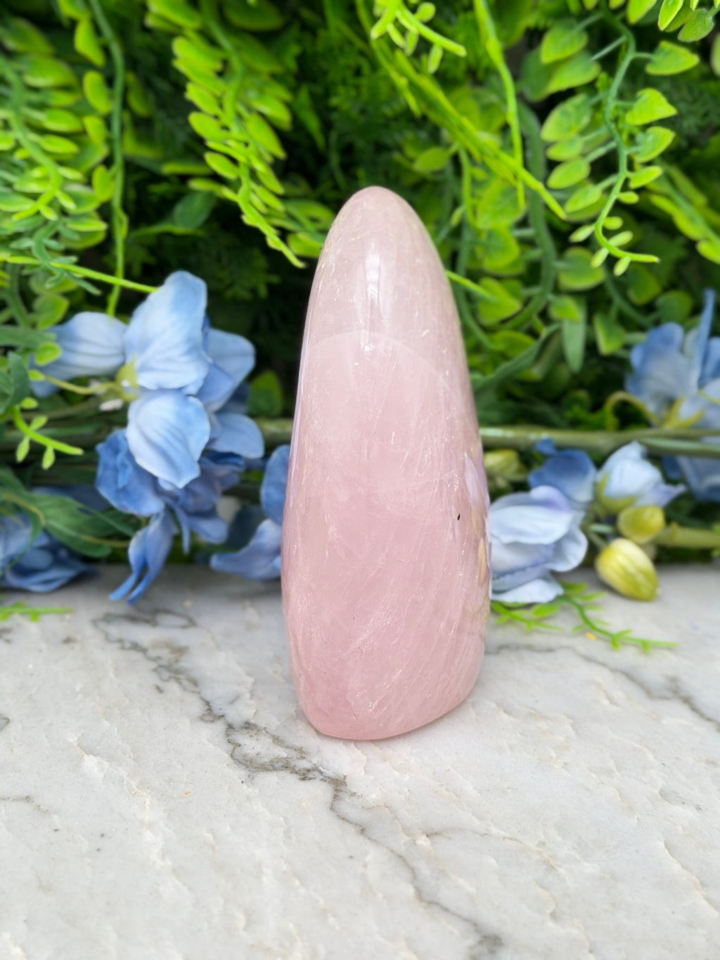 A pink crystal sits on a marble surface, surrounded by blue flowers and green foliage.