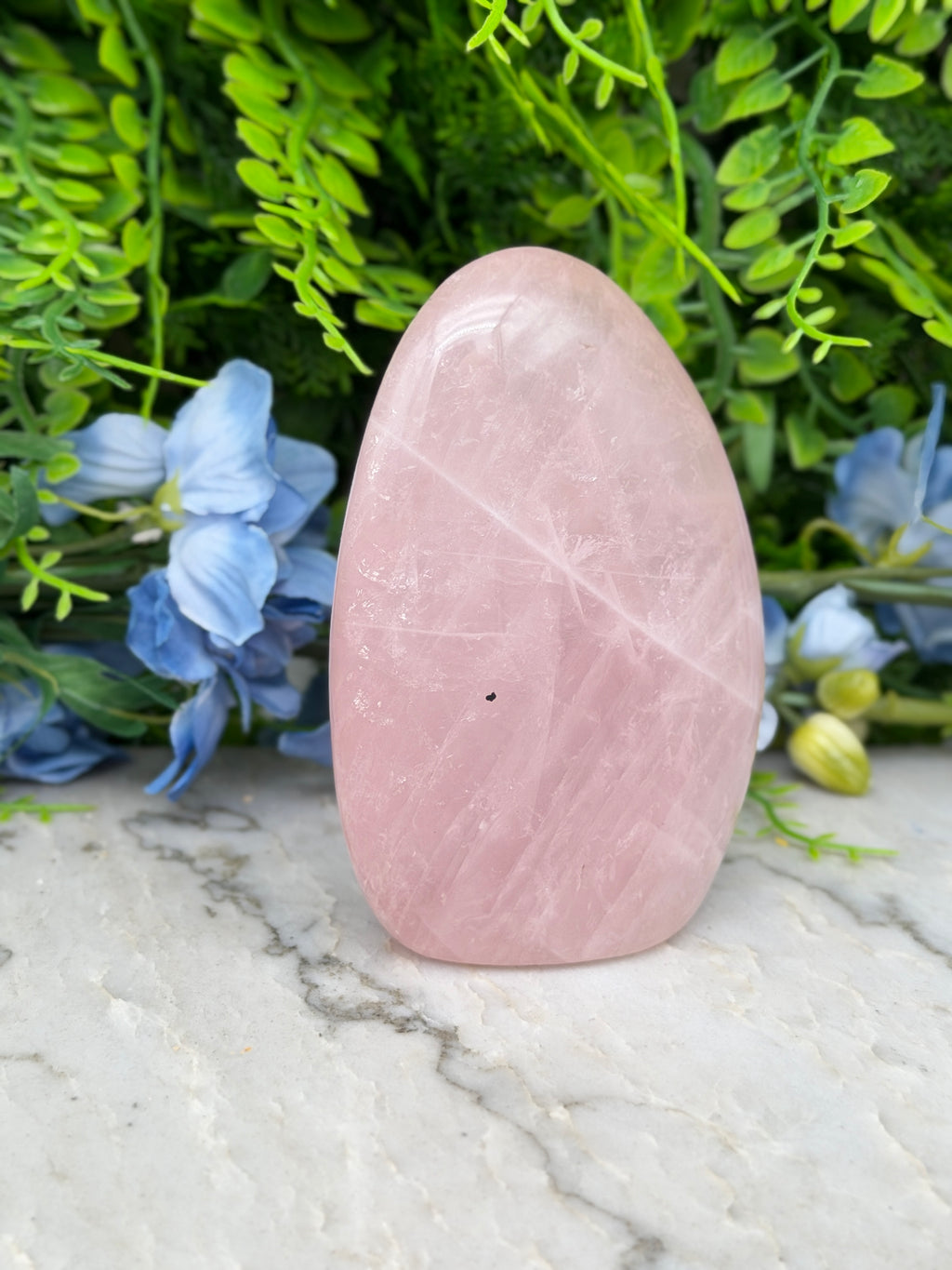 A pink crystal sits on a marble surface, surrounded by blue flowers and green foliage.