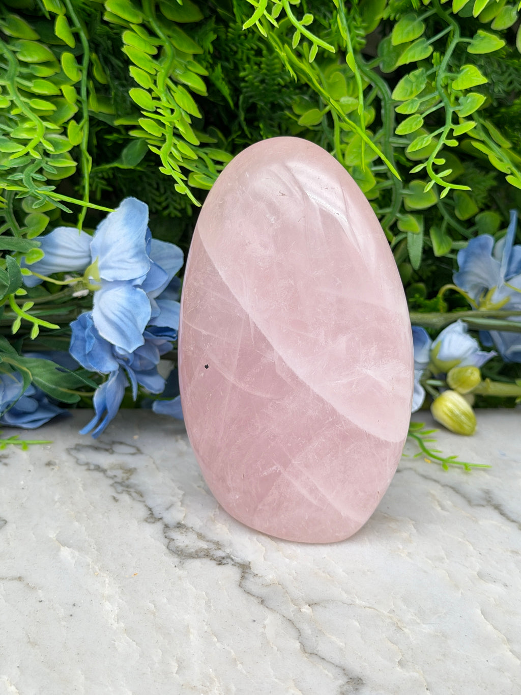 A pink crystal sits on a marble surface, surrounded by blue flowers and green foliage.