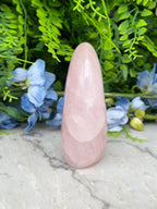 A pink crystal sits on a marble surface, surrounded by blue flowers and green foliage.