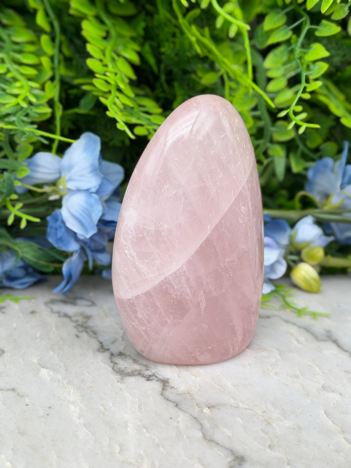 A pink crystal sits on a marble surface, surrounded by blue flowers and green foliage.