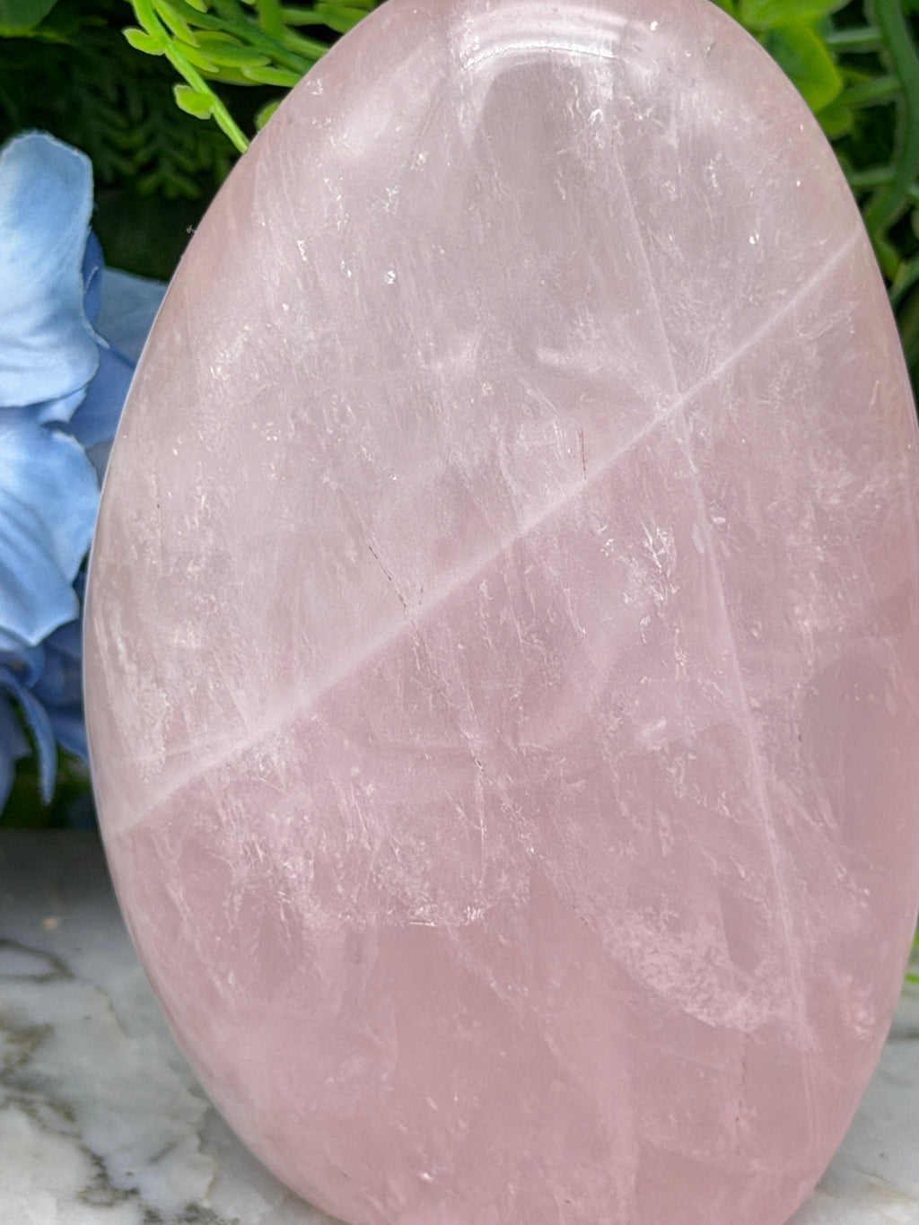A pink crystal sits on a marble surface, surrounded by green foliage.