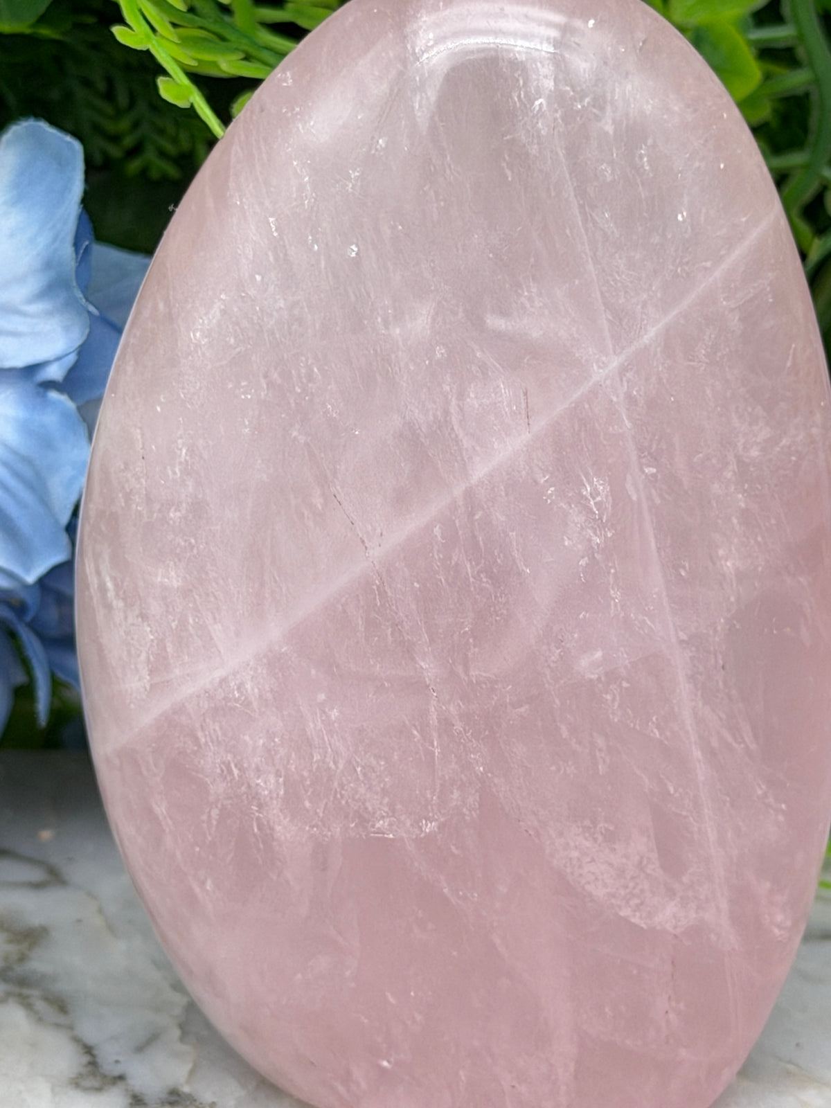 A pink crystal sits on a marble surface, surrounded by green foliage.