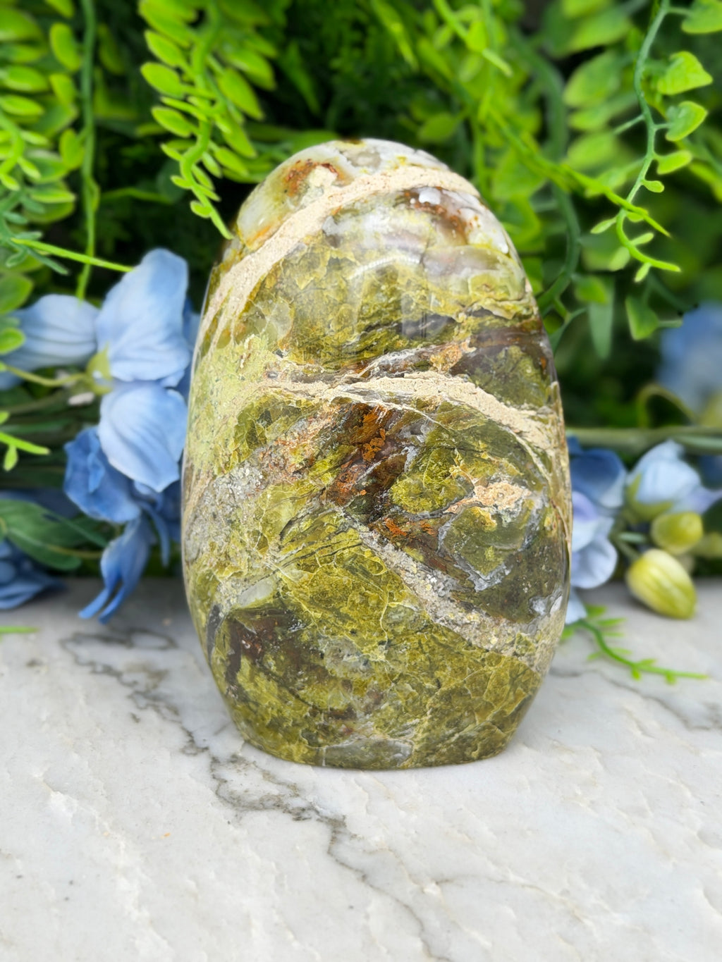 a large, jagged, and colorful stone with a mix of green, brown, and orange hues, set against a backdrop of lush greenery and blue flowers.