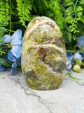 a large, jagged, and colorful stone with a mix of green, brown, and orange hues, set against a backdrop of lush greenery and blue flowers.