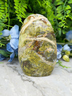 a large, jagged, and colorful stone with a mix of green, brown, and orange hues, set against a backdrop of lush greenery and blue flowers.
