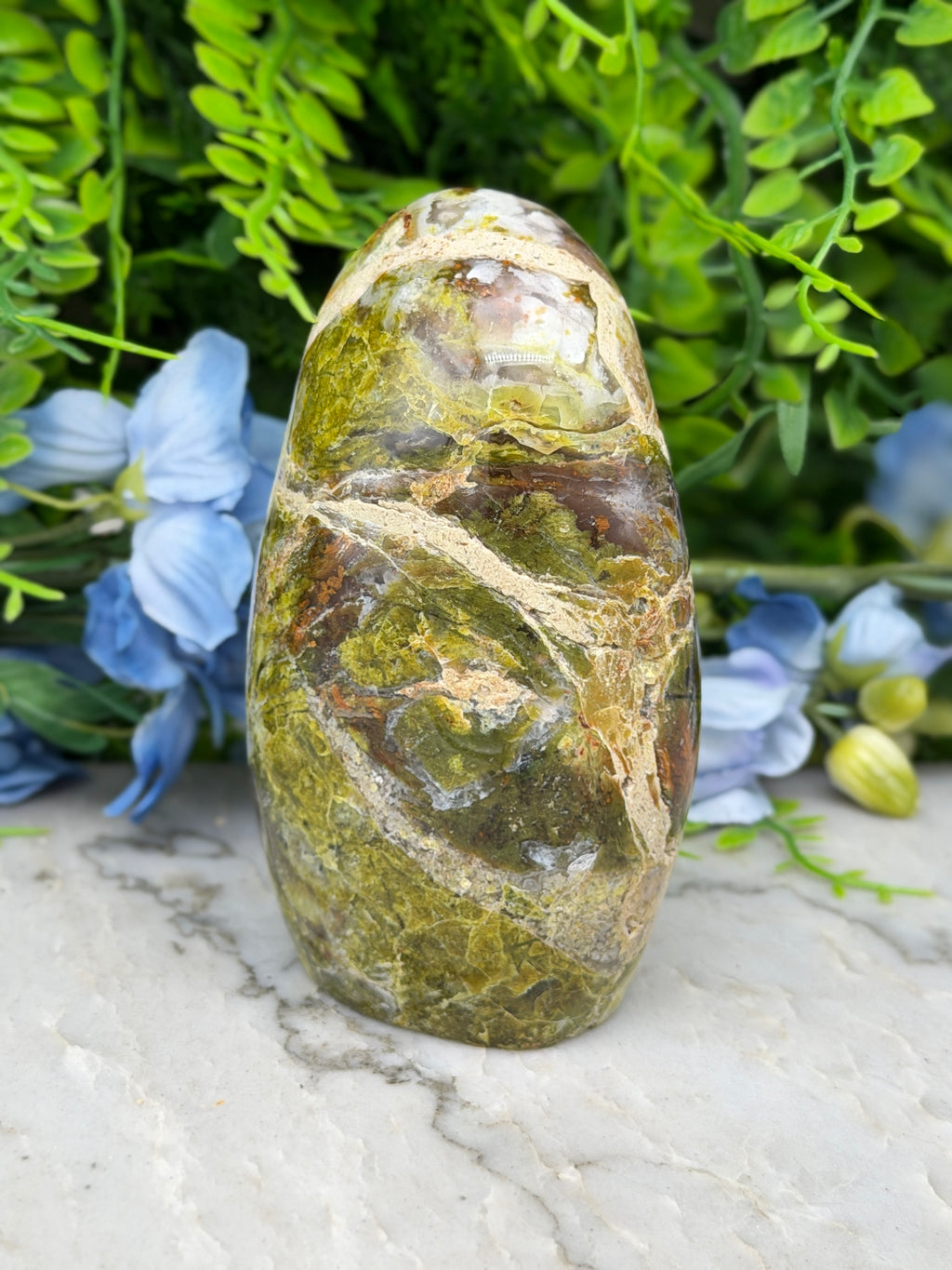 a large, green and brown rock with a unique pattern, placed on a marble surface surrounded by blue flowers.
