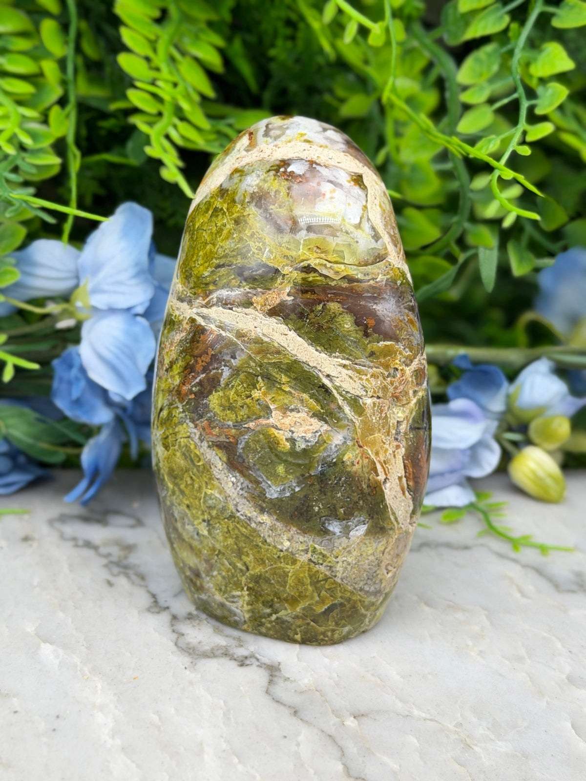 a large, green and brown rock with a unique pattern, placed on a marble surface surrounded by blue flowers.