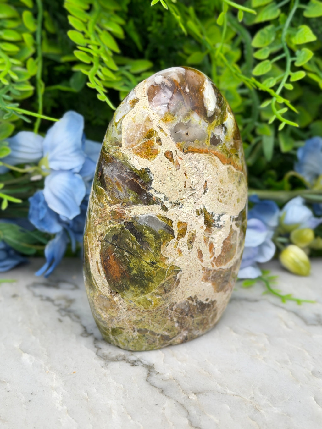 a natural stone with a unique, colorful pattern, surrounded by lush greenery and blue flowers.